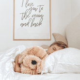 Child asleep in bed with the weighted puppy beside them, under a framed print reading I love you to the moon and back