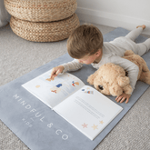 Child lying on the grey Coal mat with a plush dog, turning the pages of an open book