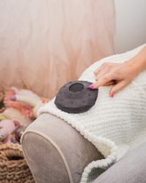 An adult's hand pressing a button on a charcoal Zenimal+ resting on a knitted throw.