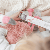 Hands holding a pink flamingo plush and a box of bath fizzies on a muslin blanket.