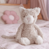 The cream weighted cat sitting upright on a bed beside a pink flower cushion