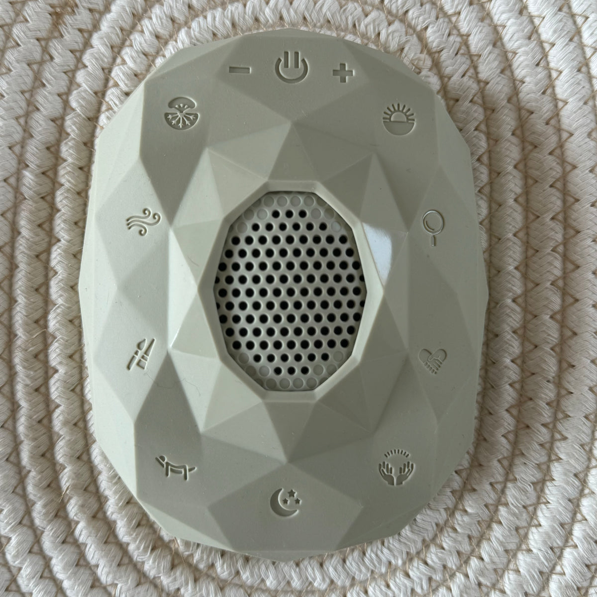 Sage gray faceted Zenimal+ viewed from above on a woven cream placemat, showing the speaker grille, power and volume buttons, and nine track icons