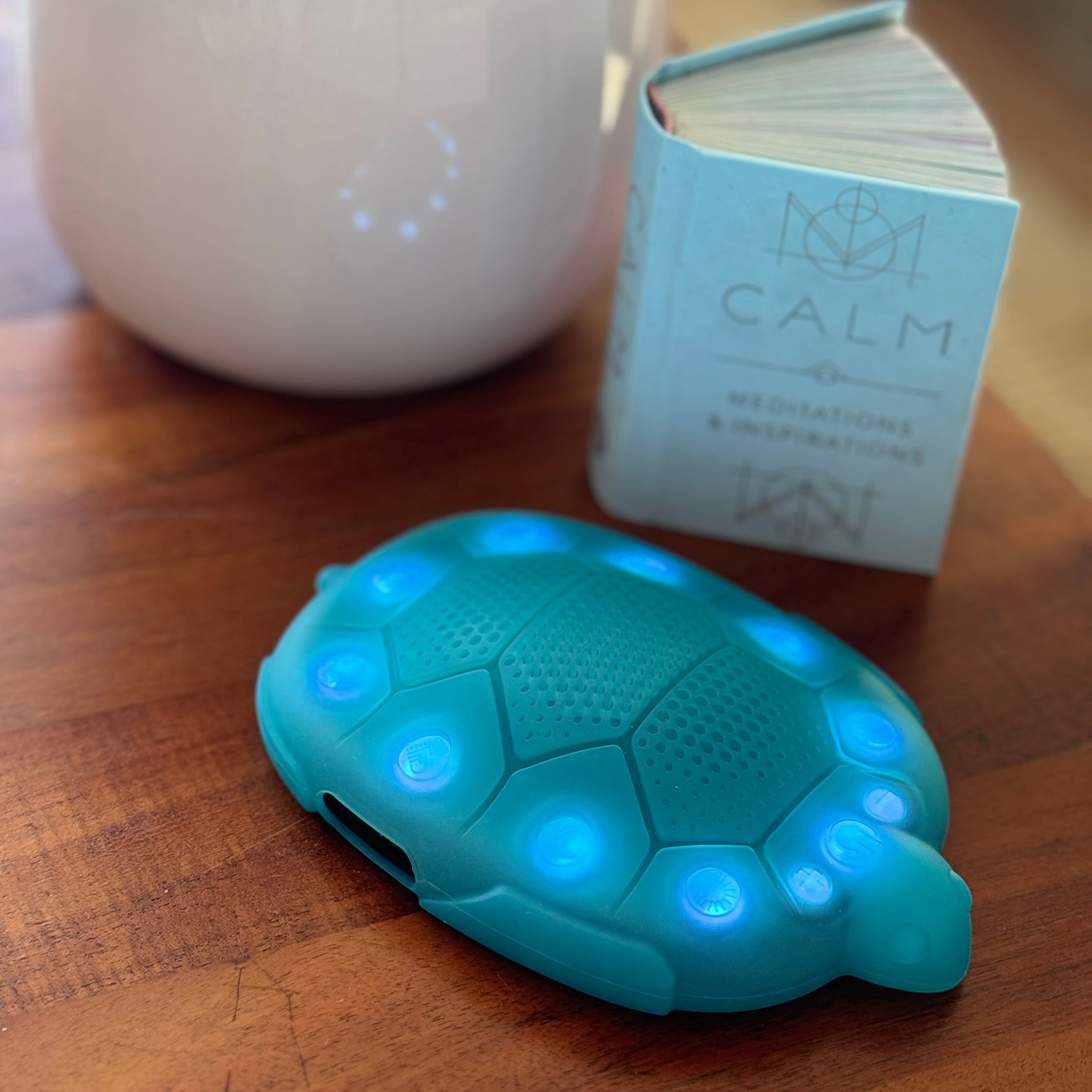Turquoise turtle-shaped device with glowing blue lights on a wooden surface, next to a white container and a small book.