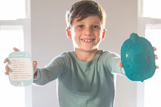 Child in light green pajamas holding two teal items on a bed.