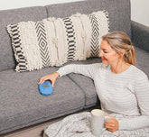 Woman sitting on the floor by a gray sofa, smiling and resting her hand on a blue Zenimal on the cushion while holding a mug