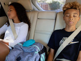 Two kids with eyes closed resting in the back seat of a car, a teal turtle Zenimal Kids sitting on a backpack between them